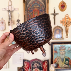 May include: A dark brown, hammered metal bowl with a rounded body and three short, tapered legs. The exterior surface features a textured, dimpled finish with copper-toned highlights visible across the uneven, metallic surface of the vessel.