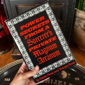 Könnte beinhalten: Ein schwarzes Buch mit einem roten Titel, der "Power Secrets From A Sorcerer's Private Magnum Arcanum" von Gavin Frost und Yvonne Frost lautet. Das Buch hat einen weißen dekorativen Rand.