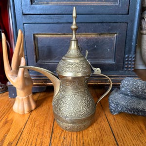 Puede incluir: Una cafetera de latón vintage con intrincados diseños florales y de hojas. La cafetera tiene un pico largo, un asa y una tapa decorativa con un remate puntiagudo. La cafetera se exhibe sobre una superficie de madera, con una escultura de mano de madera en el fondo.