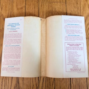 May include: An open book with the title "The Miracle of Universal Psychic Power" by A.G. Manning. The book's pages are aged, with text on the left and right pages. The cover is white with black text. The book is on a wooden surface.