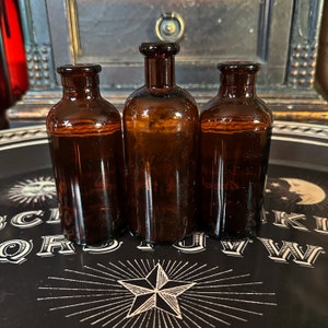 Könnte beinhalten: Drei bernsteinfarbene Glasflaschen mit Korkstopfen, die auf einer schwarzen Oberfläche mit einem weißen Stern und Buchstaben stehen.