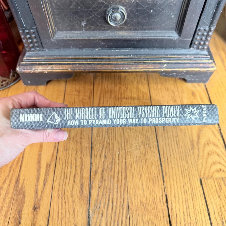 May include: A vintage book titled "The Miracle of Universal Psychic Power" by Manning. The book's spine is gray with white text and a geometric design. The book is held against a wooden floor and a dark wooden cabinet.