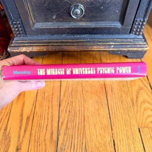 May include: A vintage book titled "THE MIRACLE OF UNIVERSAL PSYCHIC POWER" by Manning. The book has a pink cover with gold lettering and blue text. The book is held in a person's hand.