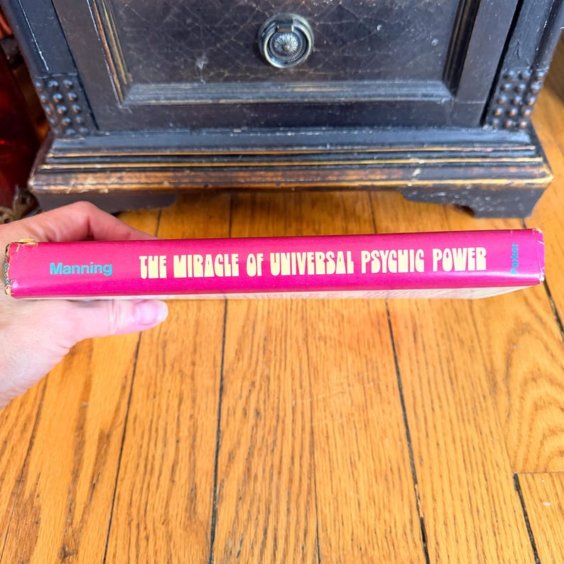 May include: A vintage book titled "THE MIRACLE OF UNIVERSAL PSYCHIC POWER" by Manning. The book has a pink cover with gold lettering and blue text. The book is held in a person's hand.
