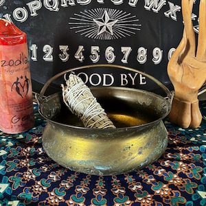 Puede incluir: Un caldero de latón con asa, sentado sobre una tela estampada. Un manojo de salvia blanca está dentro del caldero. El caldero está delante de una tabla de Ouija negra con las palabras "GOOD BYE" visibles.