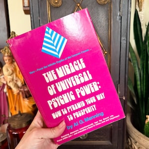 May include: A vintage book titled "The Miracle of Universal Psychic Power" by Al G. Manning. The book has a bright pink cover with white and blue text and a geometric design. The book's subtitle is "How to Pyramid Your Way to Prosperity."