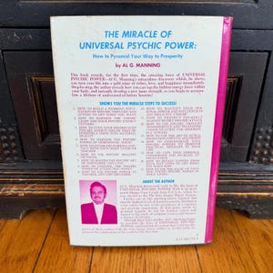 May include: The back cover of a vintage book titled "The Miracle of Universal Psychic Power" by Al G. Manning. The cover is pink and white with text detailing the book's contents and the author's biography.