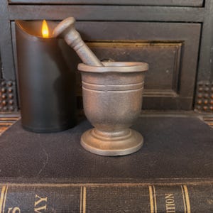 May include: A small, antique-style mortar and pestle set made of a dark, metallic material. The mortar has a rounded base and two horizontal grooves. The pestle rests inside the mortar. A black candle with a flickering flame is to the left.