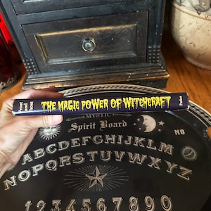 K&ouml;nnte beinhalten: Nahaufnahme eines Buches mit dem Titel "The Magic Power of Witchcraft" von Frost and Frost. Das Buch liegt auf einem schwarzen Ouija-Brett mit gelbem Alphabet und Zahlen.