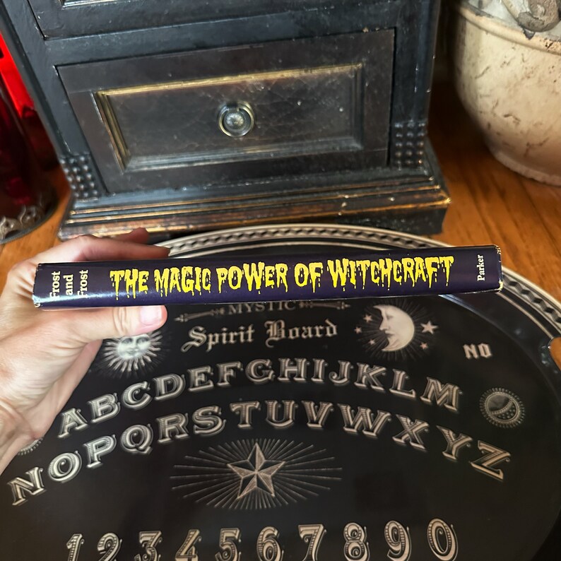 K&ouml;nnte beinhalten: Nahaufnahme eines Buches mit dem Titel "The Magic Power of Witchcraft" von Frost and Frost. Das Buch liegt auf einem schwarzen Ouija-Brett mit gelbem Alphabet und Zahlen.
