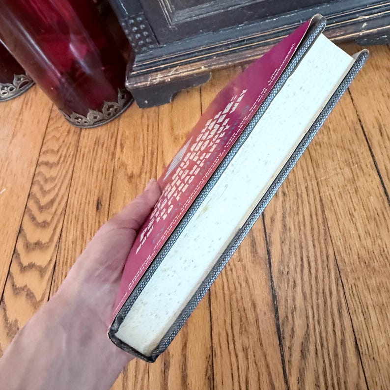 May include: A hardcover book with a red cover and white text. The book's spine is gray, and the pages are off-white. The book is held in a person's hand, with the wooden floor in the background.