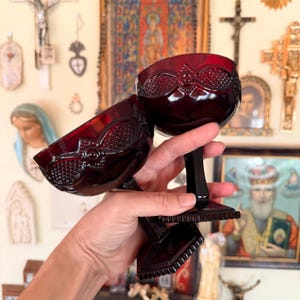 May include: Two deep red glass goblets featuring a textured diamond pattern on the bowl, a smooth stem, and a square base. The glass has a rich, dark crimson hue and a reflective surface, showcasing a repeating geometric design around the rim.