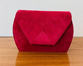 May include: A red velvet clutch with a geometric flap closure. The bag has a rounded shape and sits on a wooden surface. The clutch is a deep shade of red.