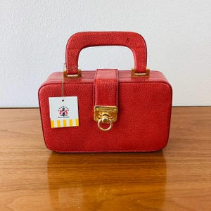 May include: A red, rectangular handbag with a top handle and gold-colored hardware. The bag has a textured surface and a clasp closure. A tag with a logo hangs from the handle.