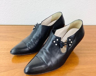 May include: A pair of black leather shoes with pointed toes. Each shoe has a small decorative cluster of pearls near the top and a small heel. The shoes are on a wooden surface.
