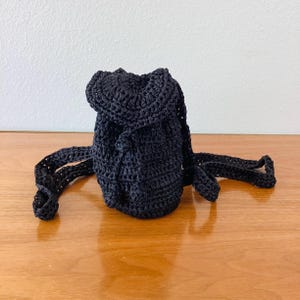 May include: A small, black crochet backpack with adjustable straps. The drawstring closure and rounded shape give it a casual, handmade look. The bag is sitting on a wooden surface.