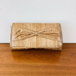 May include: A tan clutch with a bow detail. The rectangular bag is made of a snakeskin-like material. The bow is centered on the front of the bag. The bag is sitting on a wooden surface.