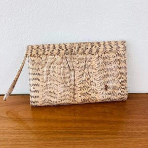May include: A beige and brown snakeskin-patterned clutch with a wrist strap. The rectangular bag has a folded design and a subtle sheen. The clutch is resting on a wooden surface.