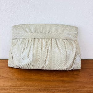 May include: A cream-colored clutch with a snakeskin pattern. The rectangular bag has a folded top and vertical seams. The clutch is made of a textured material and sits on a wooden surface.