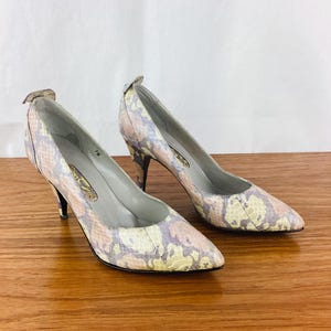 May include: A pair of pointed-toe pumps featuring a snakeskin-inspired pattern in muted shades of yellow, lavender, and tan. These shoes have a slim stiletto heel, a low-cut vamp, and a light gray interior lining with a visible size 7B label.
