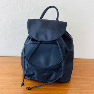 May include: A navy blue backpack with a drawstring closure and a top handle. The bag has a flap closure and a small pocket on the front. The brand name "VERSACE" is visible on the front pocket.