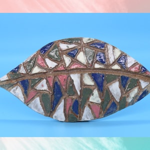 May include: A ceramic wall hanging in the shape of a leaf, featuring a mosaic design. The mosaic is made up of small, irregular pieces of ceramic in shades of blue, green, pink, and white.