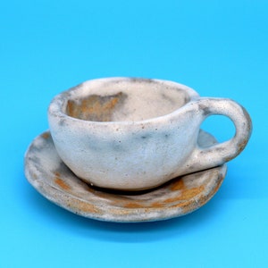 May include: A white ceramic teacup and saucer with a speckled glaze. The cup has a rounded shape and a handle. The saucer has a slightly raised edge.