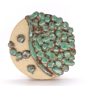 May include: A round ceramic wall hanging with a green and brown glaze. The design features a stylized face with a smile and a textured, leafy pattern.