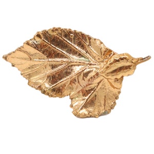 May include: A gold leaf brooch with a detailed, realistic design. The leaf is slightly curled at the edges, giving it a natural look.