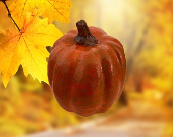 May include: A red and orange pumpkin with a stem, against a blurred background of fall foliage.