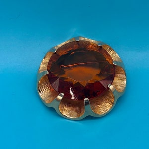 Könnte beinhalten: Eine goldfarbene Brosche mit einem großen, facettierten, bernsteinfarbenen Glasstein in der Mitte. Die Brosche ist blumenförmig mit gewellten Rändern.