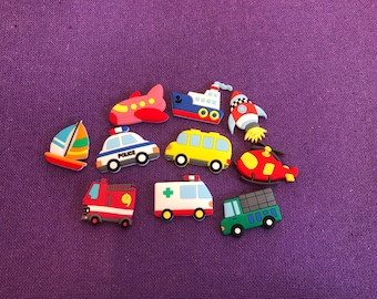 May include: A collection of colorful transportation-themed charms, including a sailboat, airplane, police car, school bus, fire truck, ambulance, helicopter, ship, rocket, and a green truck, all on a purple background.