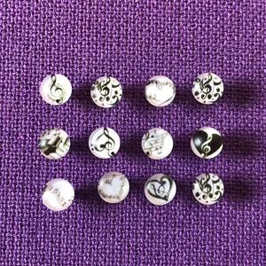 May include: Twelve white round buttons with black musical notes and hearts printed on them. The buttons are arranged in three rows of four on a purple fabric background.