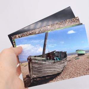 May include: A set of postcards featuring a weathered wooden boat on a beach with a blue sky and white clouds. The boat is partially submerged in the sand and has a green hull. There is another smaller boat in the background.