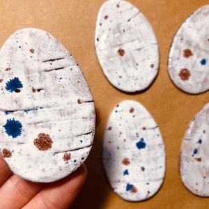 May include: Five white ceramic oval-shaped coasters with brown and blue speckles. The coasters have a rustic, handmade look.