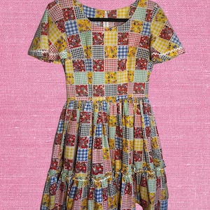 May include: A vibrant patchwork dress with short sleeves and a square neckline. The dress showcases a mix of patterns, including gingham and floral prints in red, yellow, blue, and green. The skirt has a ruffled hemline.