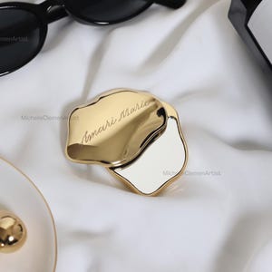 May include: A gold-toned, irregular-shaped compact mirror with a polished finish. The lid features the engraved name Amari Marie in a cursive script. The mirror is partially open, revealing a reflective surface, and is surrounded by black sunglasses.