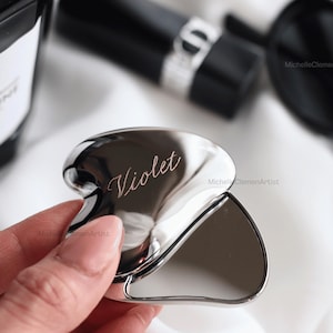 May include: A polished silver-toned heart-shaped compact mirror with the name Violet engraved in a cursive script on the front surface. The metallic object features a smooth, reflective finish and a curved, ergonomic design for handheld use.