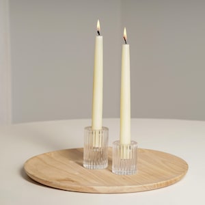 Op de afbeelding: Twee witte kaarsen in heldere glazen kaarsenhouders op een houten dienblad. De kaarsen zijn aangestoken en de vlammen zijn zichtbaar.