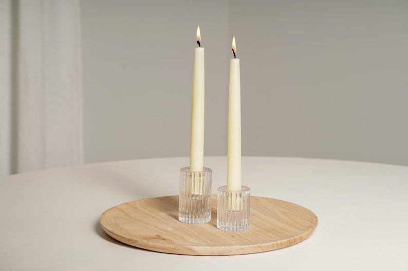 Op de afbeelding: Twee witte kaarsen in heldere glazen kaarsenhouders op een houten dienblad. De kaarsen zijn aangestoken en de vlammen zijn zichtbaar.