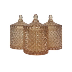 Pode incluir: Três frascos de vidro para velas com tampas. Os frascos são de cor pêssego claro e têm um padrão de diamante. As tampas têm um pequeno botão na parte superior.