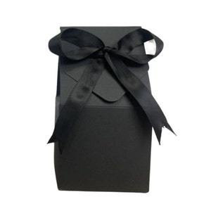 May include: A black gift box with a black satin ribbon tied in a bow. The box is made of sturdy cardboard and has a flap on the top.