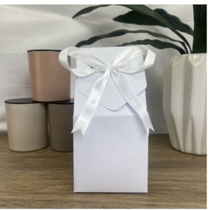 May include: A white gift box with a white satin ribbon tied in a bow. The box is sitting on a wooden surface.