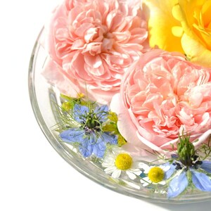 Op de afbeelding: Een glazen kom gevuld met water en verschillende bloemen, waaronder roze rozen, gele rozen en blauwe bloemen.