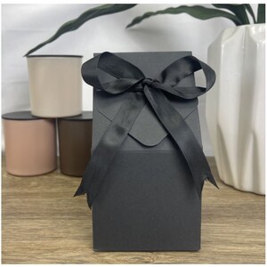 May include: A black gift box with a black satin ribbon tied in a bow. The box is sitting on a wooden surface.