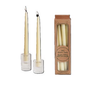 Op de afbeelding: Twee witte bijenwas kaarsen in glazen kaarsenstandaards. Een doos met 100% bijenwas kaarsen met de tekst "100% Beeswax Tapers" en "Bougies Effil&eacute;es 100% cire d'abeille" op de voorkant.