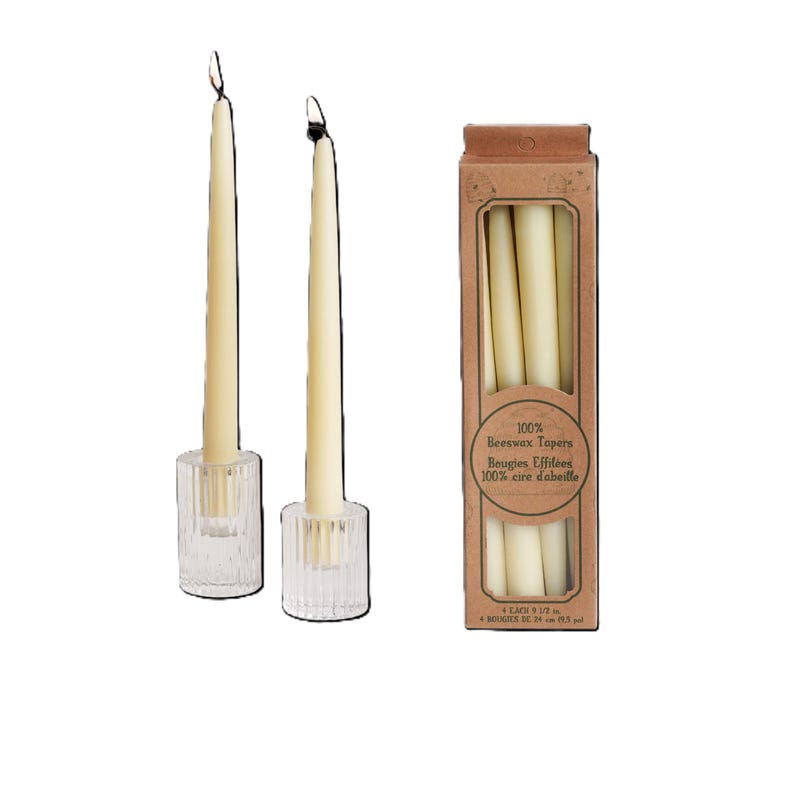 Op de afbeelding: Twee witte bijenwas kaarsen in glazen kaarsenstandaards. Een doos met 100% bijenwas kaarsen met de tekst "100% Beeswax Tapers" en "Bougies Effil&eacute;es 100% cire d'abeille" op de voorkant.