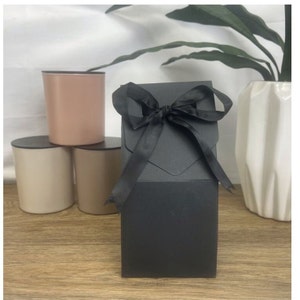 May include: A black gift box with a black satin ribbon bow. The box is sitting on a wooden surface in front of three candles.