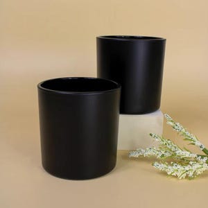 Op de afbeelding: Twee zwarte cilindrische containers. De containers zijn gemaakt van een mat materiaal en hebben een gladde, afgeronde vorm. Eén container staat iets hoger op een klein, vierkant, gebroken wit blok. Een takje witte bloemen staat op de voorgrond.