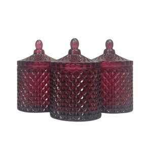 May include: Three burgundy glass candle jars with diamond-shaped patterns and lids. The jars are arranged in a row.